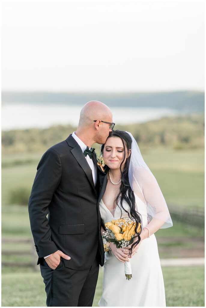 groom kissing the top of bride's head as they stand in field with susquehanna river behind them at lauxmont farms