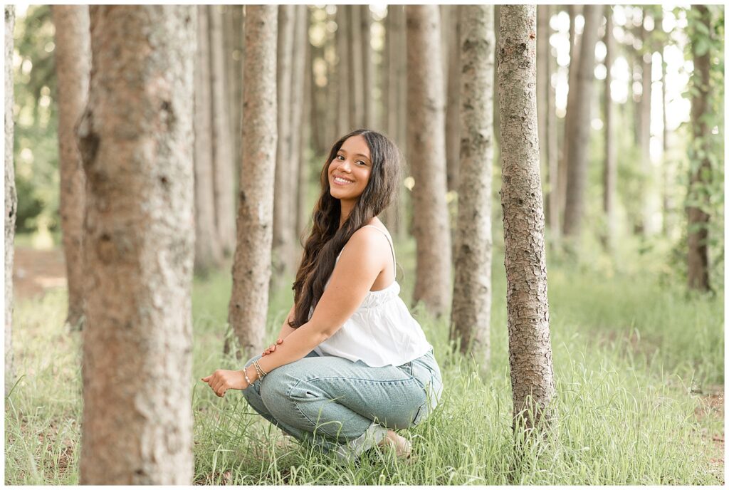 senior girl squatting down with left shoulder towards camera by rows of evergreen trees at overlook park in lancaster county
