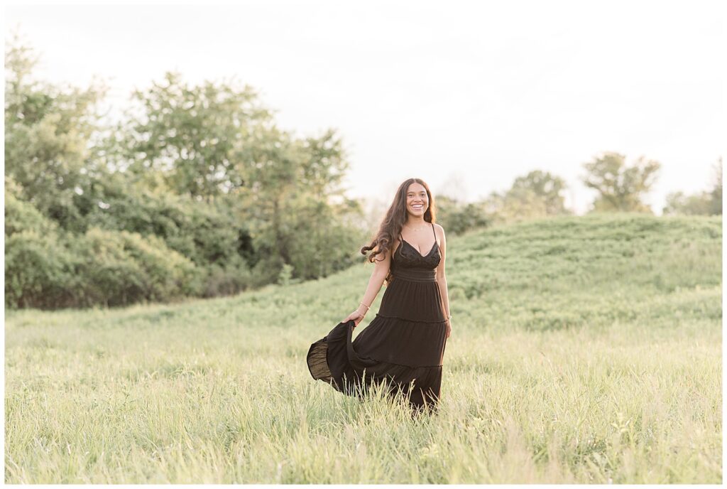 senior girl in black dress holding edge of dress in grass field at sunset in lancaster county