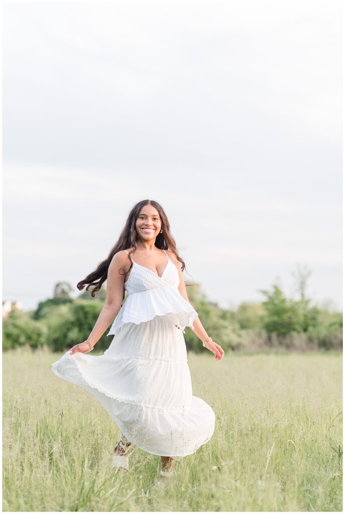 senior girl in long white dress holding edge of dress in field at park in manheim township