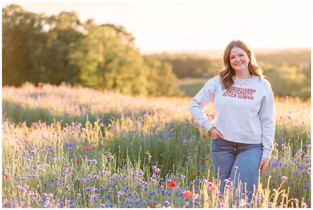 senior girl wearing susquehanna university sweatshirt and blue jeans with hand on hip at sunset for this wildflower field senior session