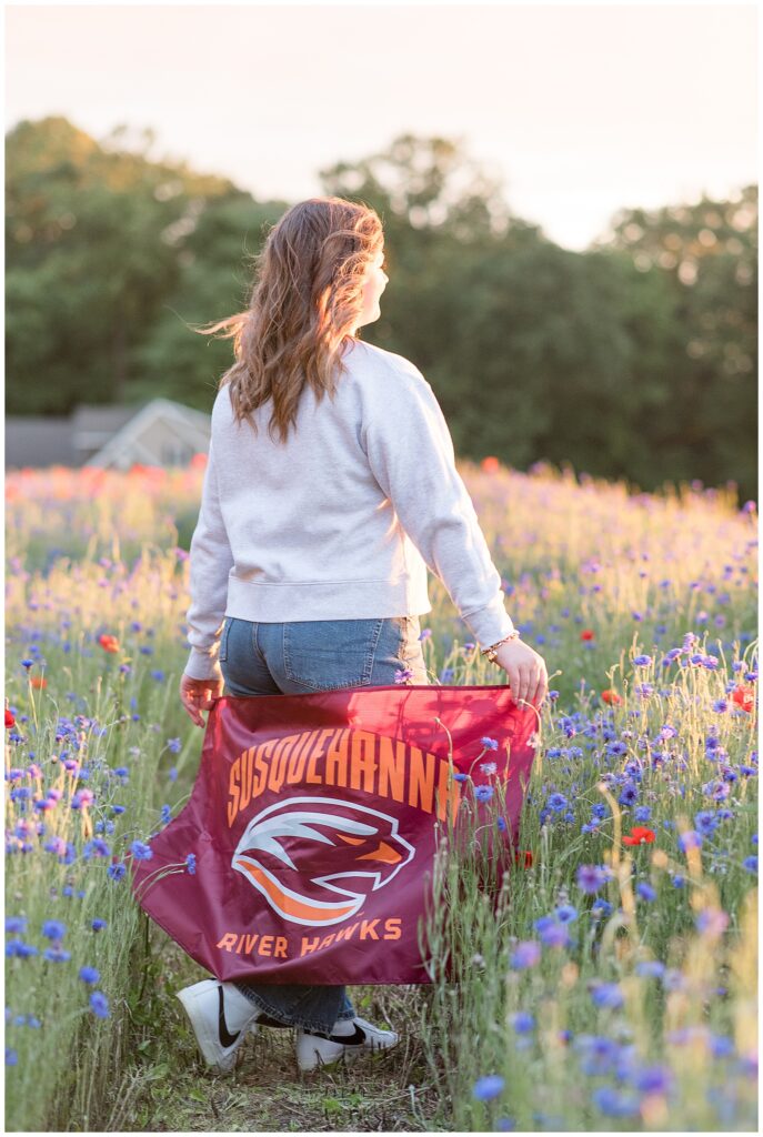 senior girl holding susquehanna university flag behind her with her back to the camera for this wildflower field senior session