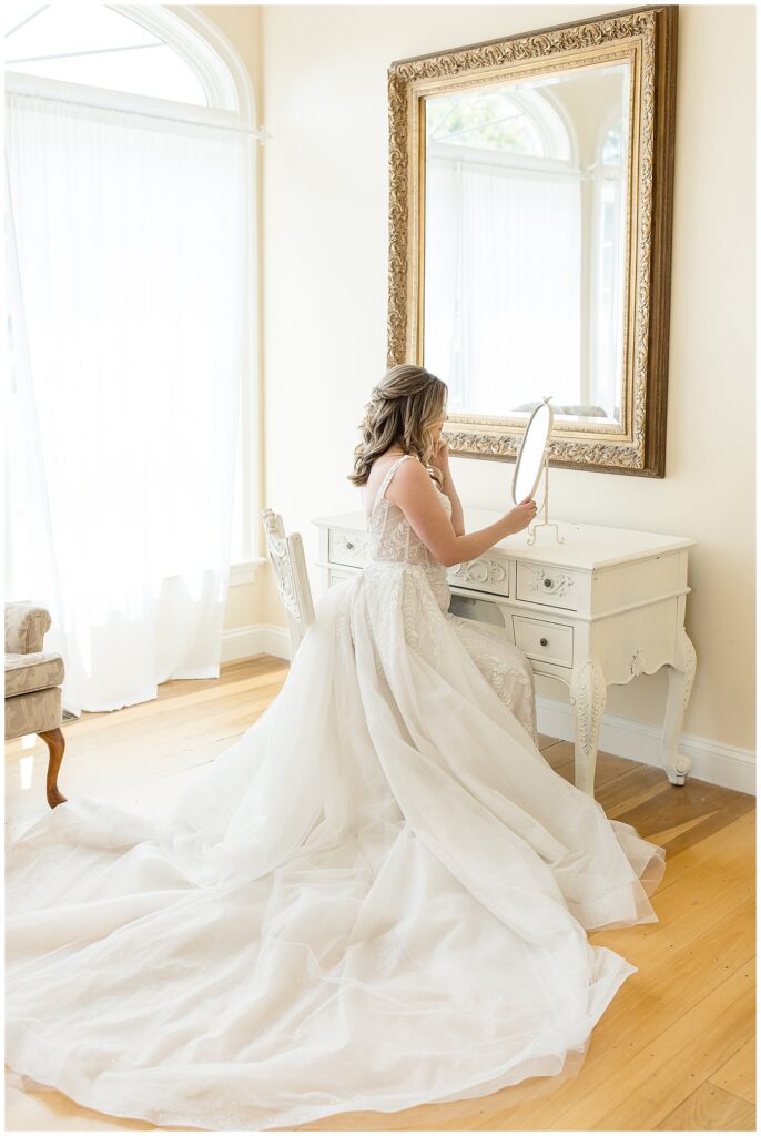 bride sitting at white desk in bridal suite looking into mirror with her long dress train flared out in landenberg pennsylvania
