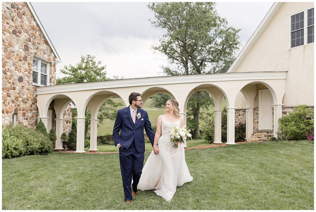 couple holding hands walking towards camera with arched walkway behind them at bryn awel manor in pennsylvania