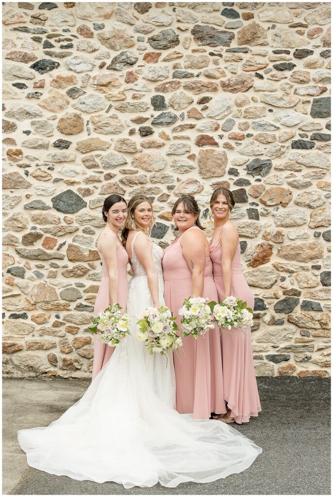 bride in white gown with her three bridesmaids in pastel pink gowns by stone wall at this Bryn Awel Manor Summer Wedding
