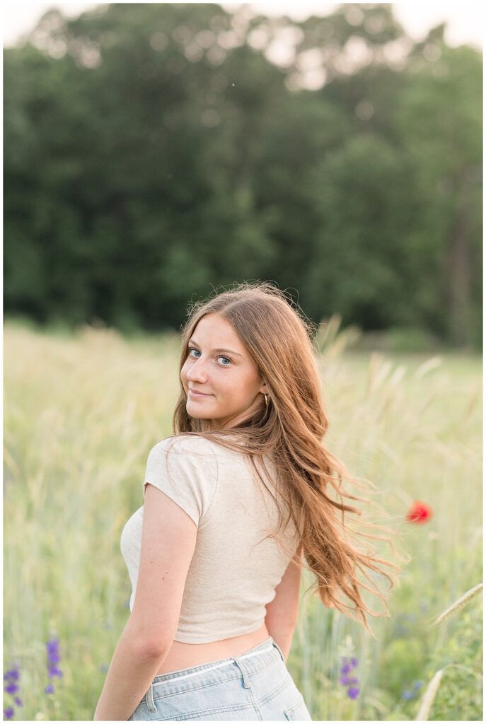 senior girl looking back over left shoulder wearing crop top and blue jeans for this wildflower field summer senior session