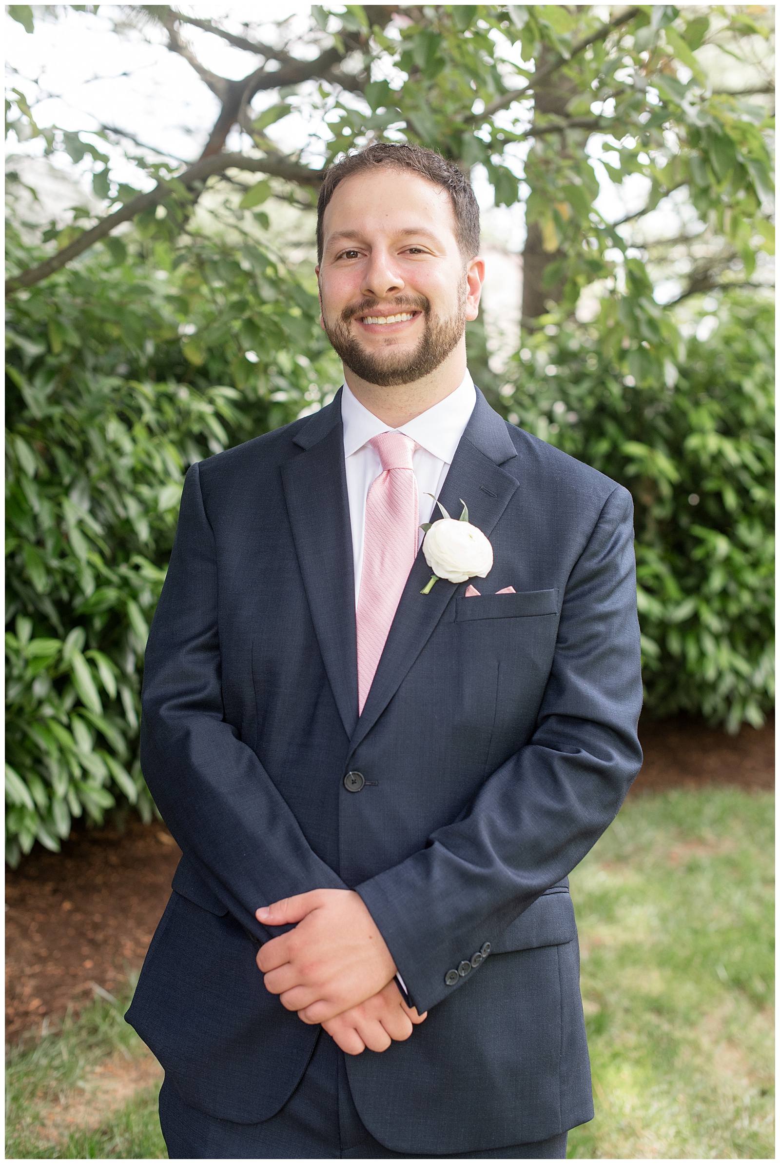 groom wearing navy blue suit with pastel pink tie smiling for camera outside at this Inn at Leola Village Wedding
