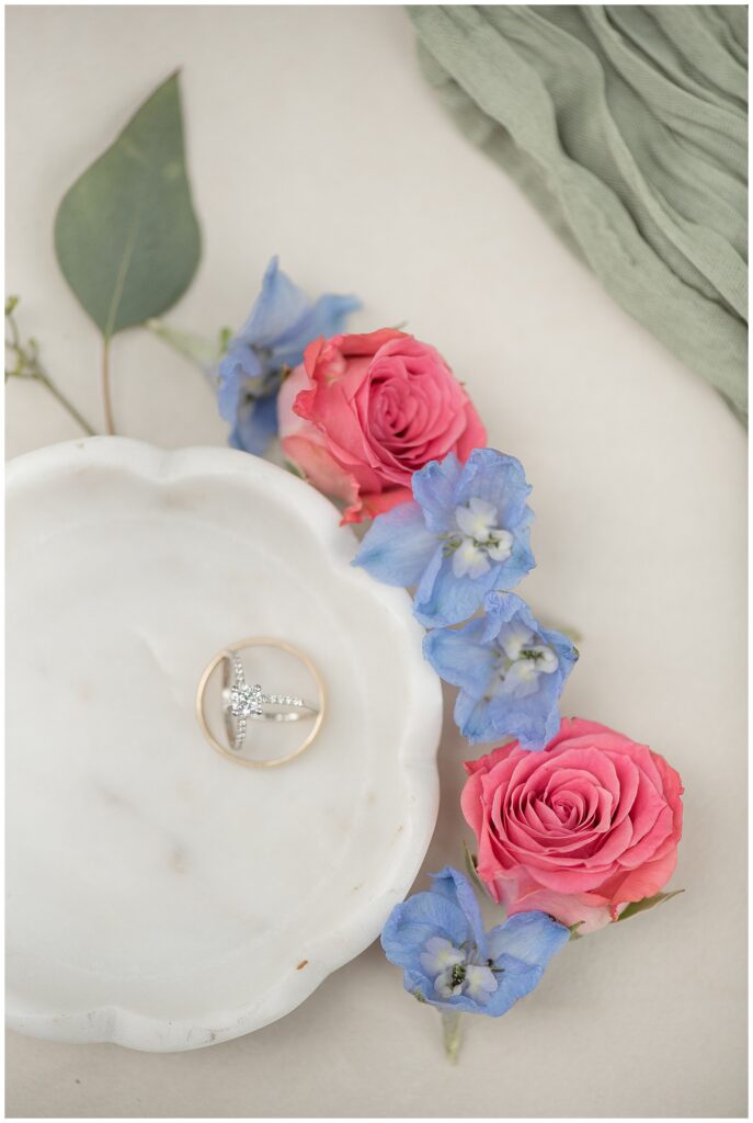 gold wedding rings displayed in scalloped marble bowl with pink roses and blue flowers along edge