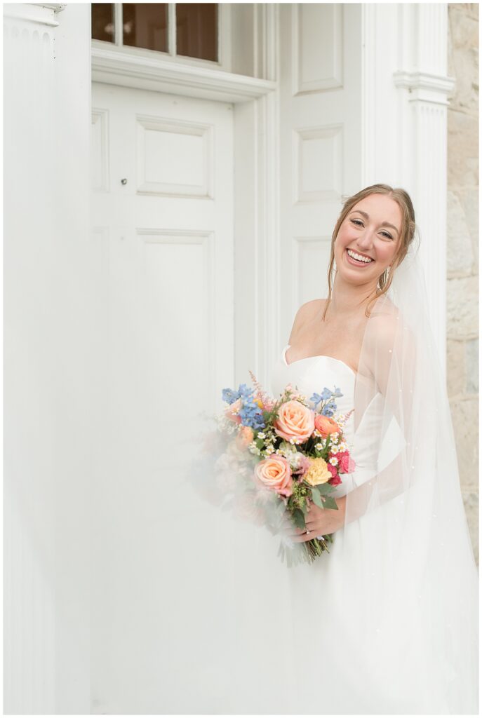 bride in white strapless gown and long veil holding colorful pink and blue bouquet by white front door of the barn at silverstone