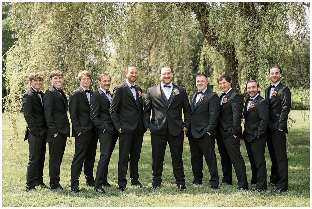 groom surrounded by his nine groomsmen all in black suits by willow tree at the barn at silverstone in lancaster pennsylvania