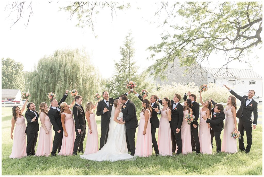 couple kissing as their entire bridal party cheers on sunny day for this Barn at Silverstone Pink and Black Wedding