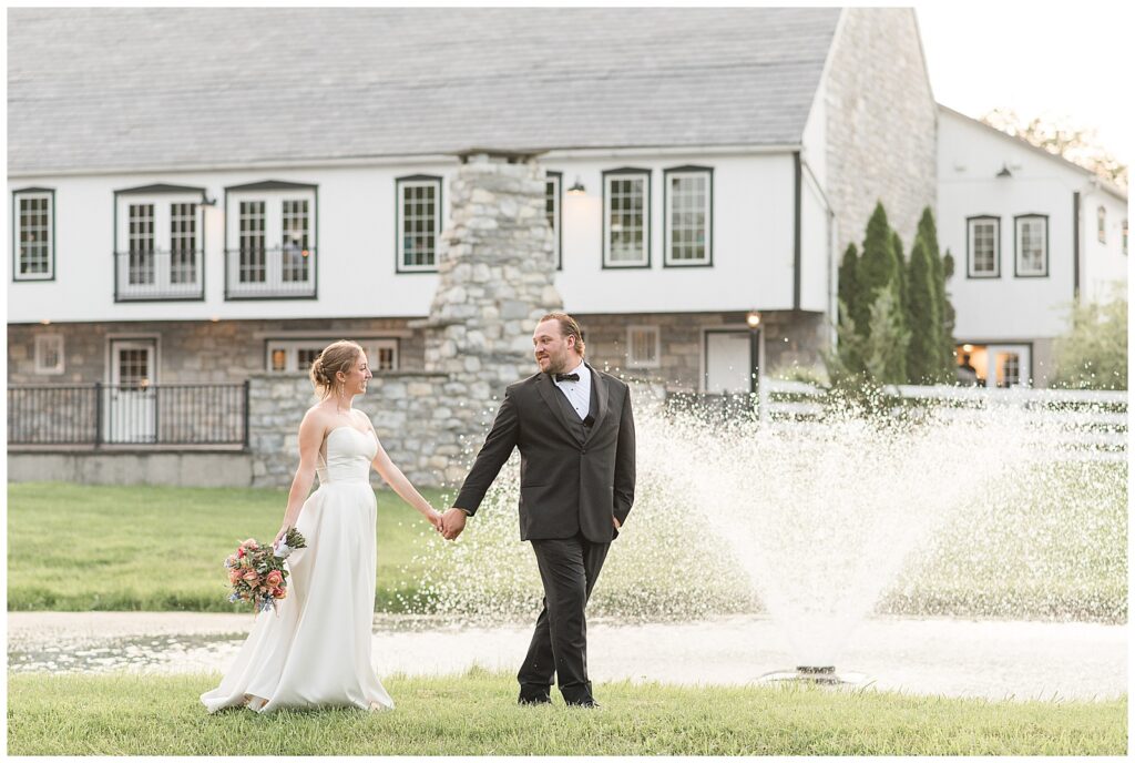 couple holding hands and walking along edge of pond with barn in background for this Barn at Silverstone Pink and Black Wedding