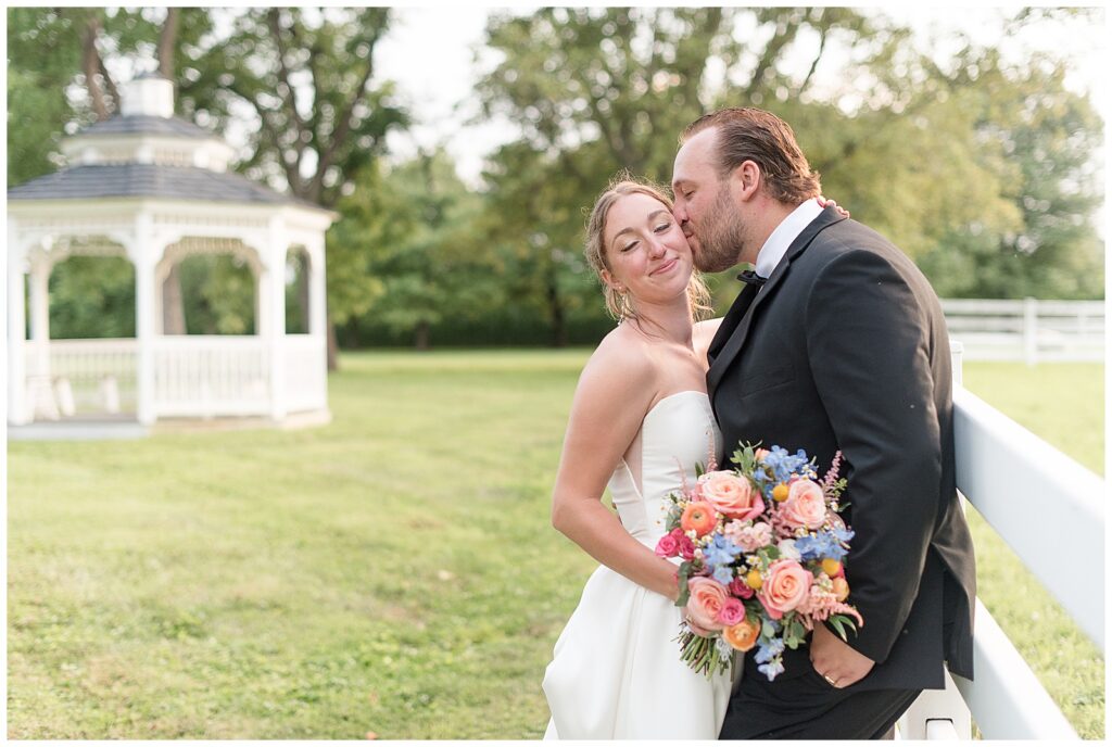 groom kissing bride's left cheek as bride holds colorful bouquet at this Barn at Silverstone Pink and Black Wedding
