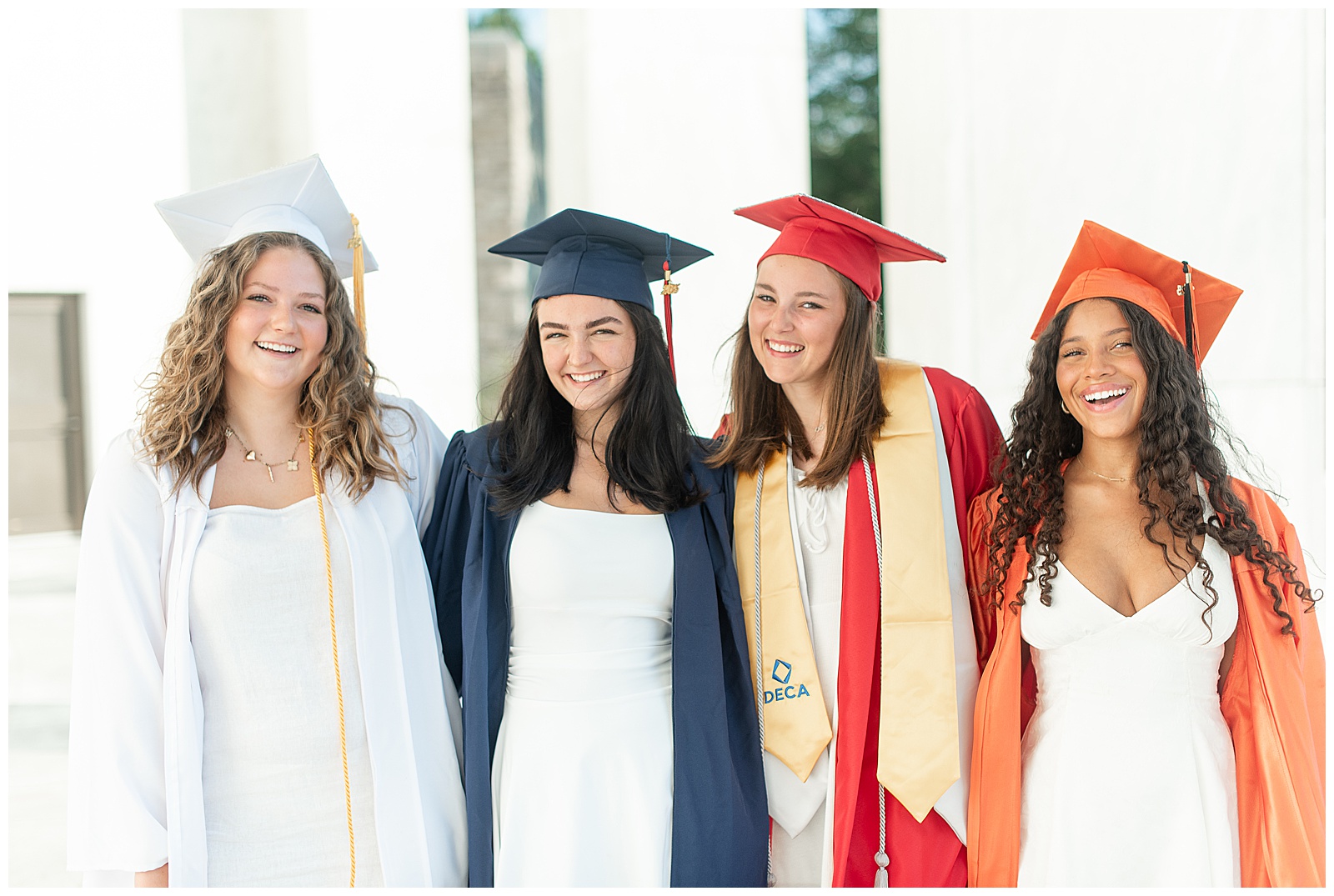 four senior girls hugging in white, navy blue, red, and orange high school graduation gowns for this Founders Hall Cap and Gown Session