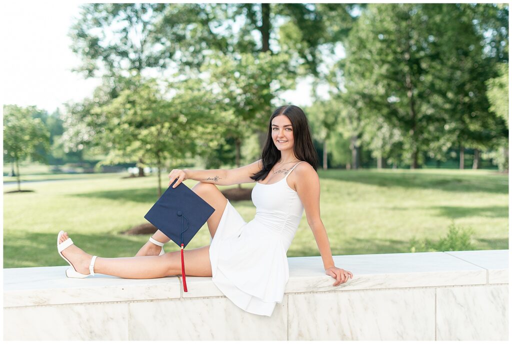 senior girl in cute white dress sitting on concrete ledge and holding her navy blue graduation cap in hershey pennsylvania at founders hall