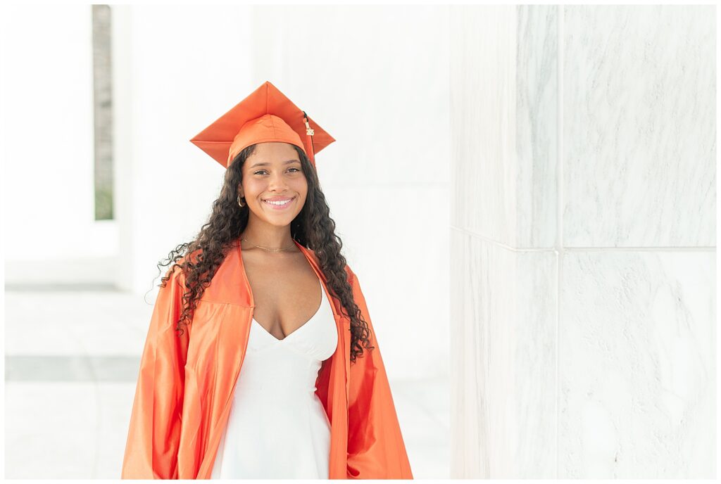 senior girl in white dress with orange graduation gown overtop by white columns at founders hall at milton hershey school