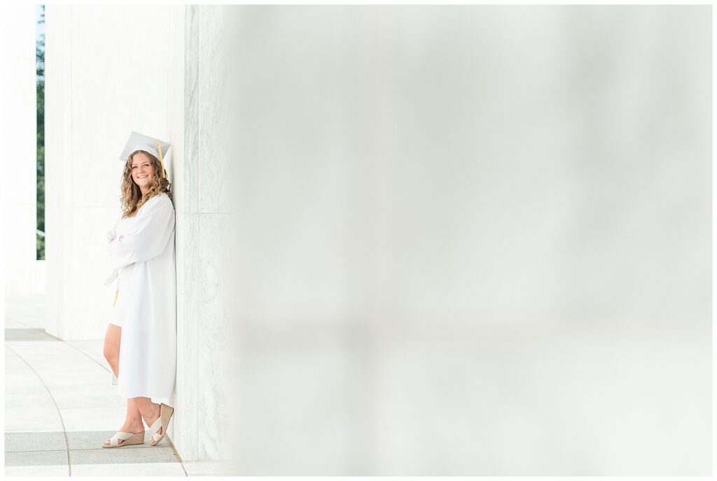 senior girl in white graduation cap and gown leaning back against wall of founders hall at milton hershey school in hershey pennsylvania
