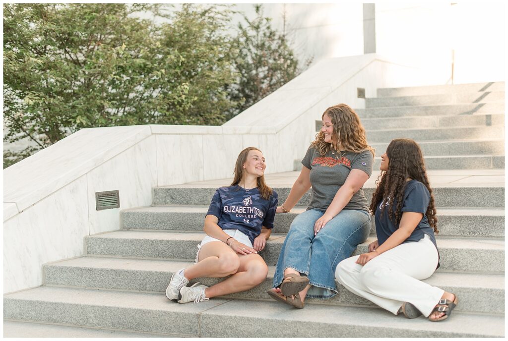 three senior girls sitting on steps wearing college t-shirts and jeans or shorts for this Founders Hall Cap and Gown Session