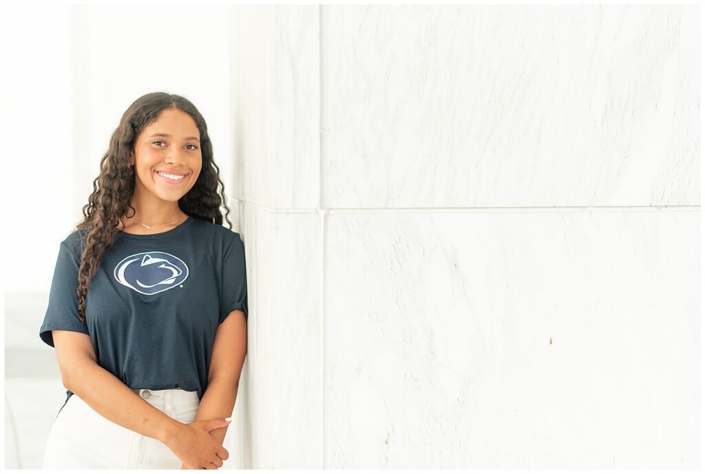senior girl wearing penn state nittany lions t-shirt leaning against white wall for this Founders Hall Cap and Gown Session