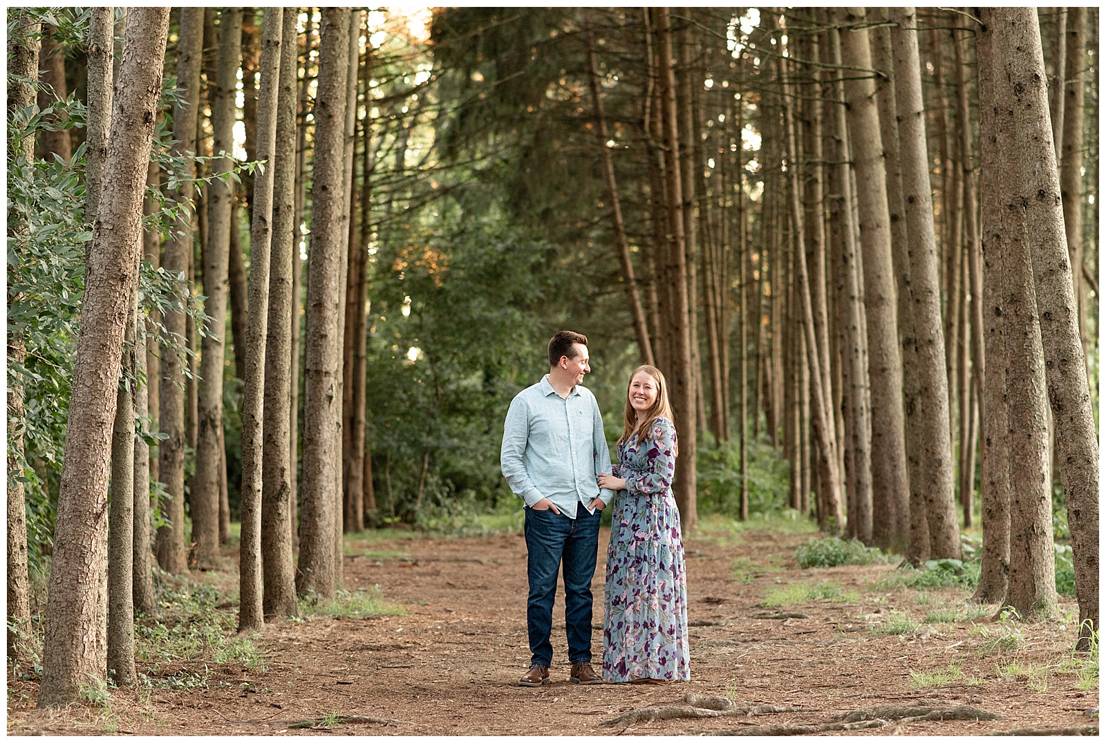 guy smiling at girl as she smiles at camera by rows of evergreen trees at this Puppy-Loving Overlook Park Engagement Session
