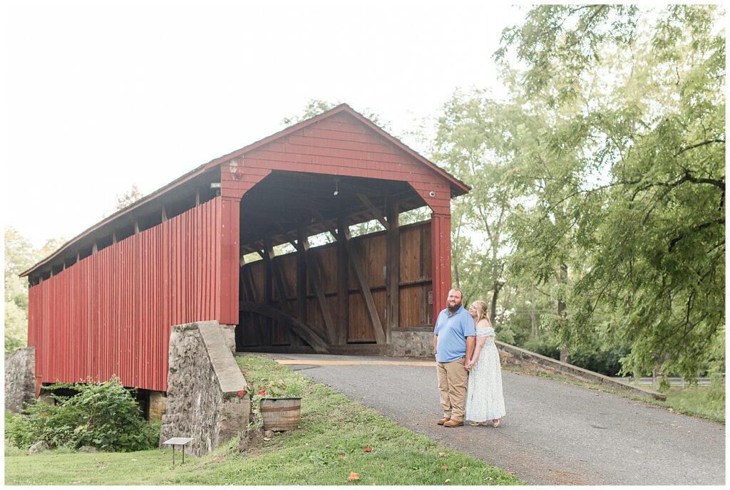woman standing behind man in front of historic red covered bridge at this Historic Poole Forge Summer Engagement Session