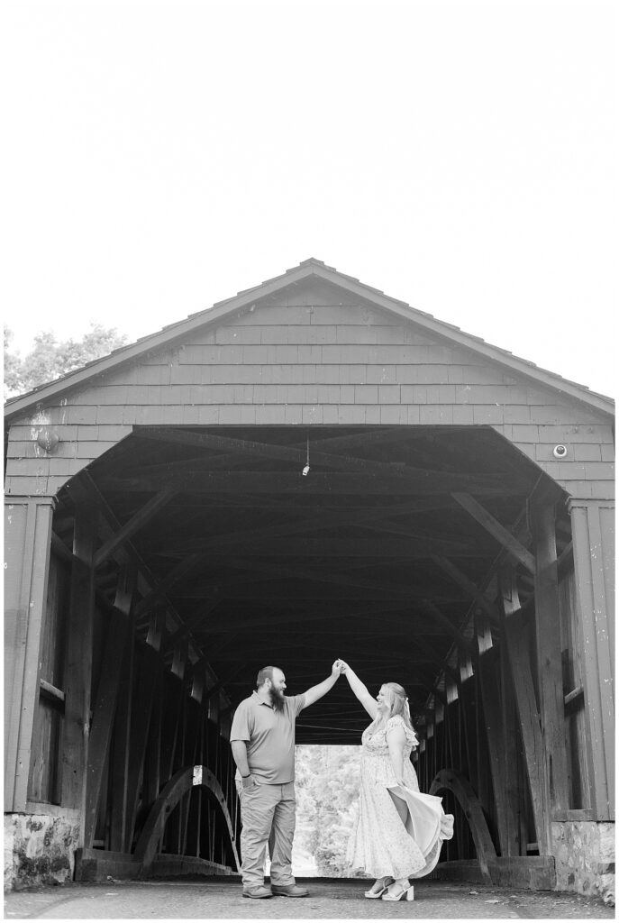 black and white photo of guy twirling girl in front of covered bridge for this Historic Poole Forge Summer Engagement Session