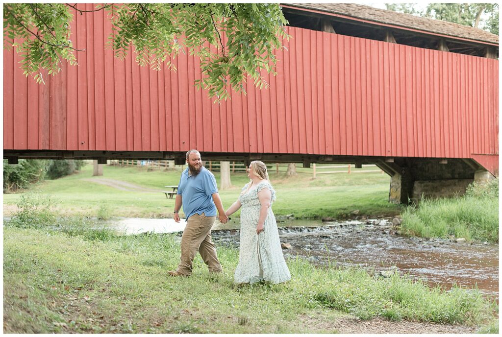 couple holding hands and walking along grass by red covered bridge for this Historic Poole Forge Summer Engagement Session
