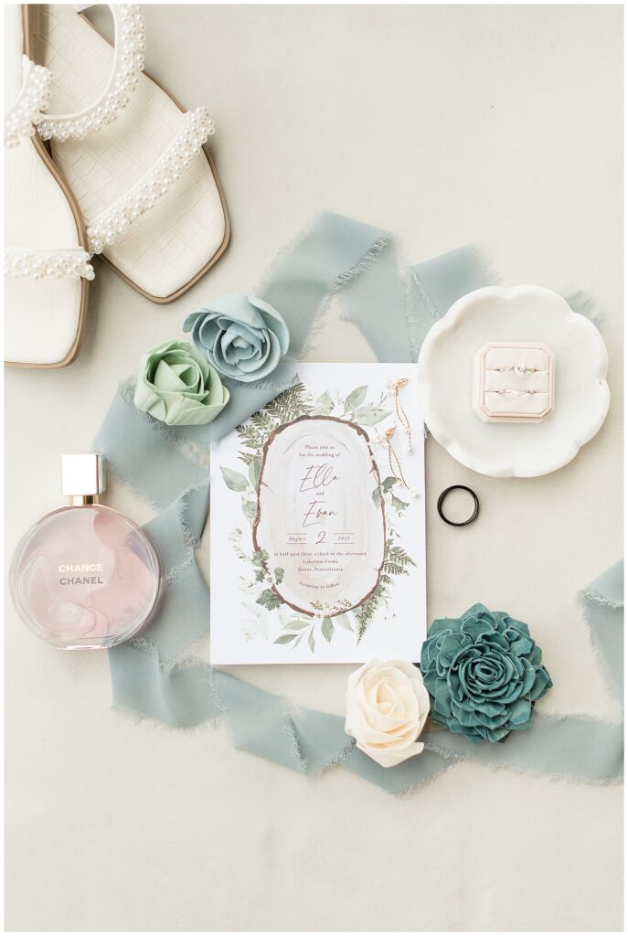 wedding invitation surrounded by blue ribbon, flowers, bride's sandals, and rings at this Lakeview Farms Summer Wedding