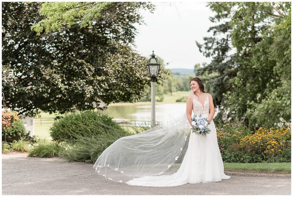 bride looking over right shoulder with her long white veil blowing in breeze behind her for this Lakeview Farms Summer Wedding
