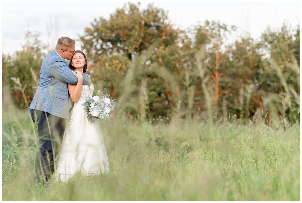 groom hugging and kissing his bride from behind in field of wild grasses at this Lakeview Farms Summer Wedding