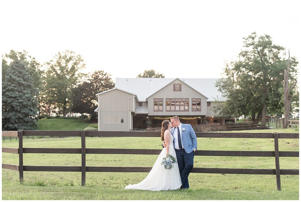 couple kissing by fence in front of venue on summer evening at this Lakeview Farms Summer Wedding