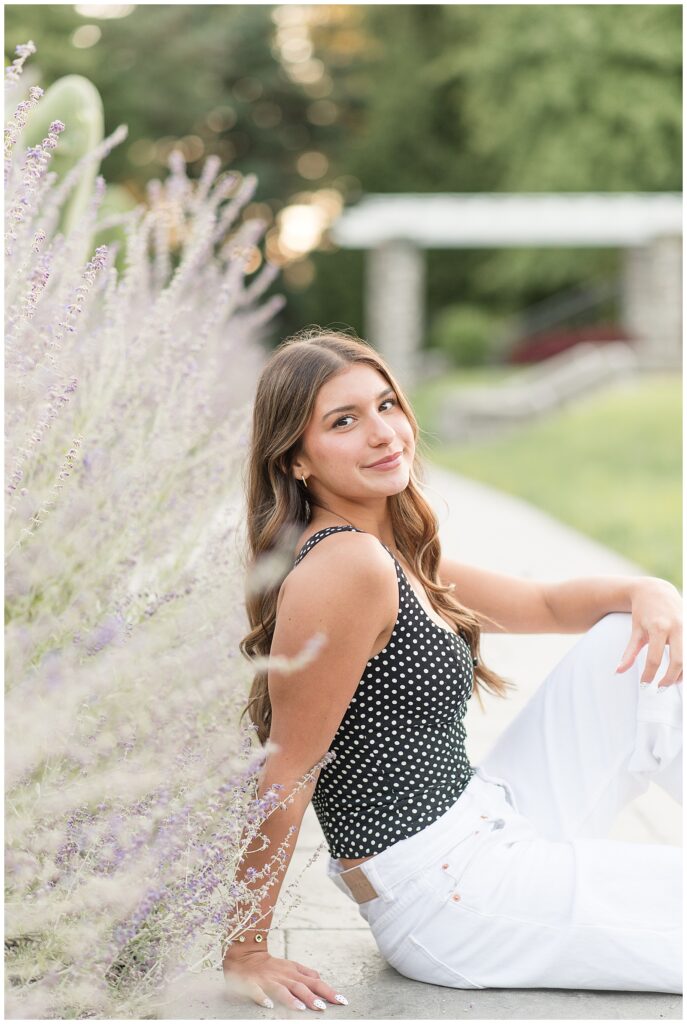 senior girl sitting on sidewalk by bush with right shoulder toward camera for this Summer Masonic Village Senior Session