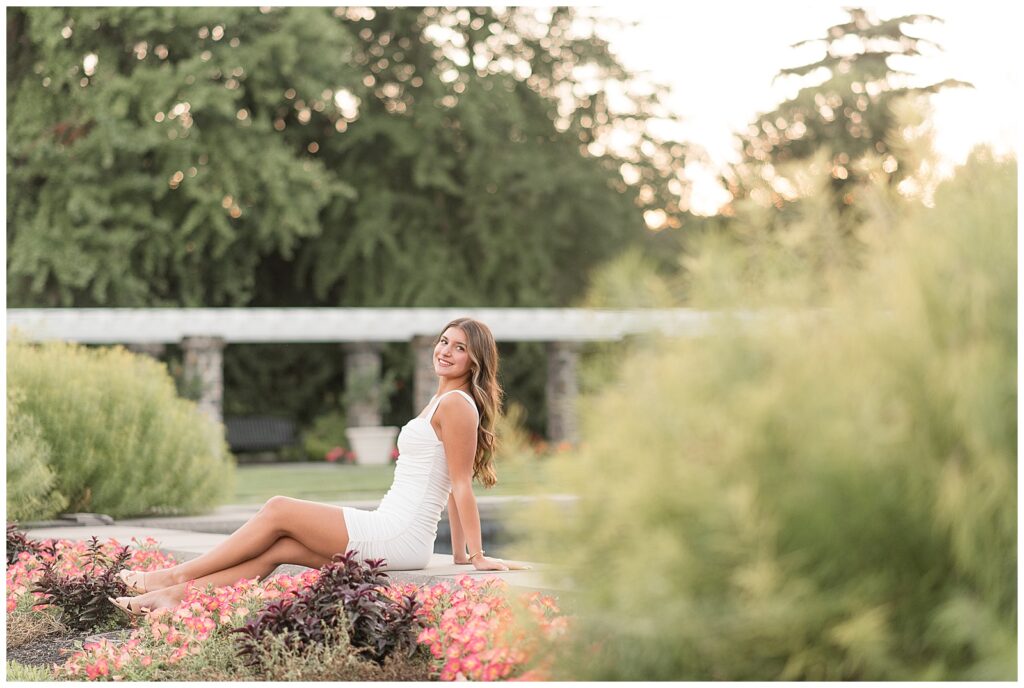 senior girl leaning back along edge of fountain in white dress for this Summer Masonic Village Senior Session