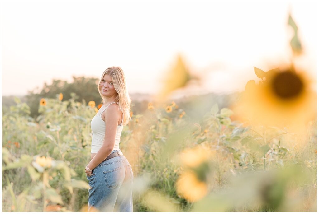 senior girl look back over left shoulder in white tank top and blue jeans for this Summer Wildflower Field Senior Session