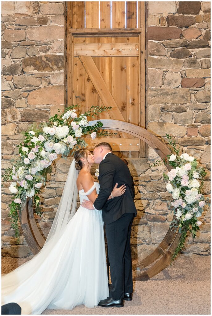 couple sharing their first kiss by round floral display for indoor wedding ceremony at elizabeth furnace