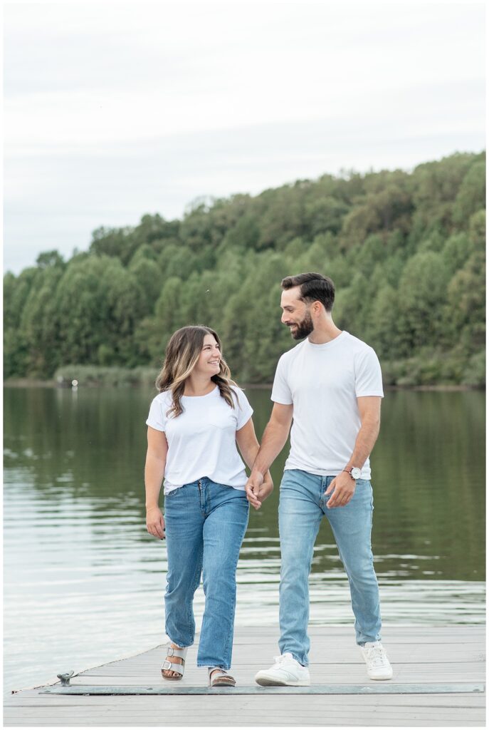 couple in white t-shirts and blue jeans holding hands and walking on dock for this Fall Hibernia Park Engagement Session