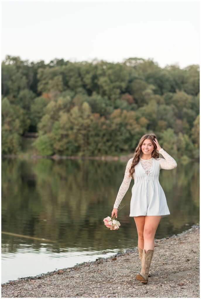 senior girl in cute white dress holding bouquet and walking along lake for this Hibernia Park Senior Session