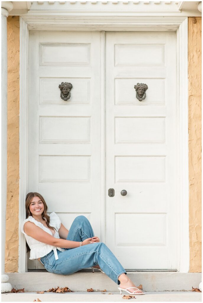 senior girl in white tank top and blue jeans leaning against white door frame of house at this Hibernia Park Senior Session