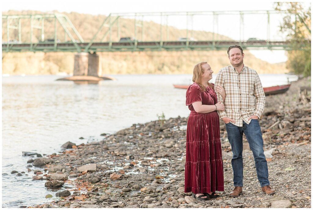 woman holding onto man's right arm as he smiles at camera by river for this Downtown New Hope Engagement Session