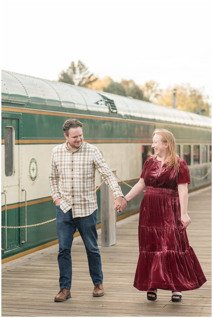 couple holding hands and walking along sidewalk beside old green train for this Downtown New Hope Engagement Session