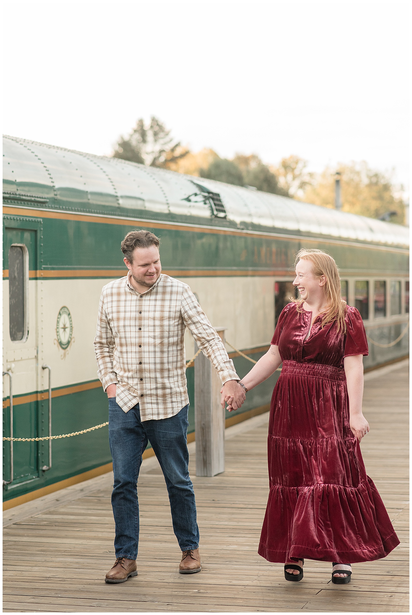 couple holding hands and walking along sidewalk beside old green train for this Downtown New Hope Engagement Session