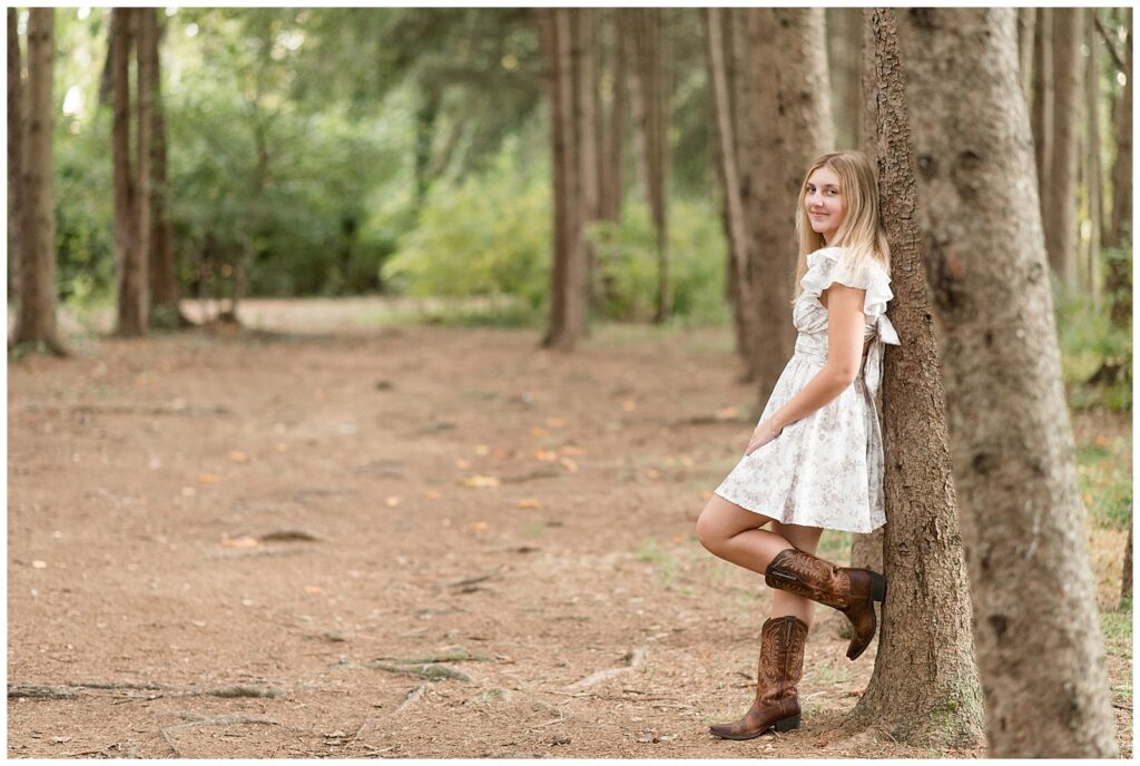 senior girl leaning against evergreen tree with left boot propped back on tree for this Fall Overlook Park Senior Session
