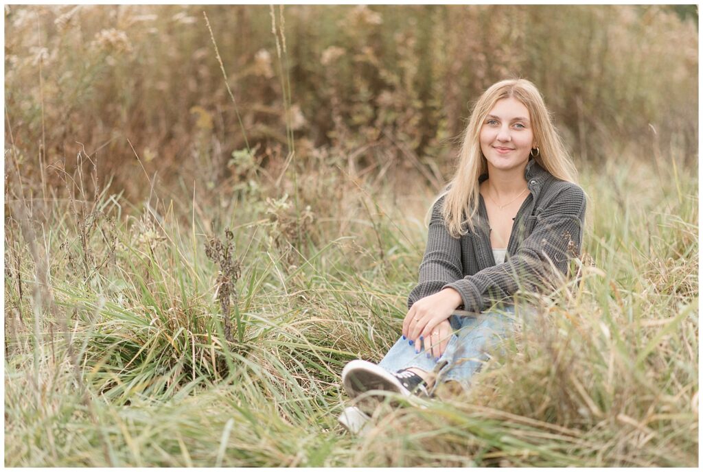 senior girl in oversized gray shirt and blue jeans sitting in wildflower field for this Fall Overlook Park Senior Session