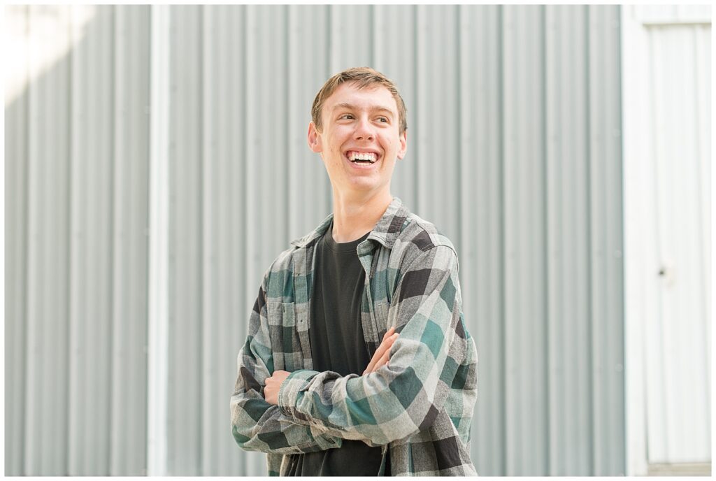 senior guy in flannel shirt with arms folded and smiling by gray barn for this Family Farm Senior Guy Session in Lancaster County