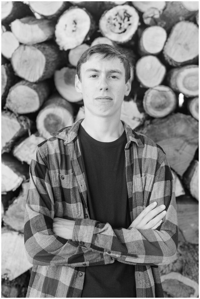 black and white photo of senior guy with arms folded in front of stacked logs for this Family Farm Senior Guy Session in Lancaster County