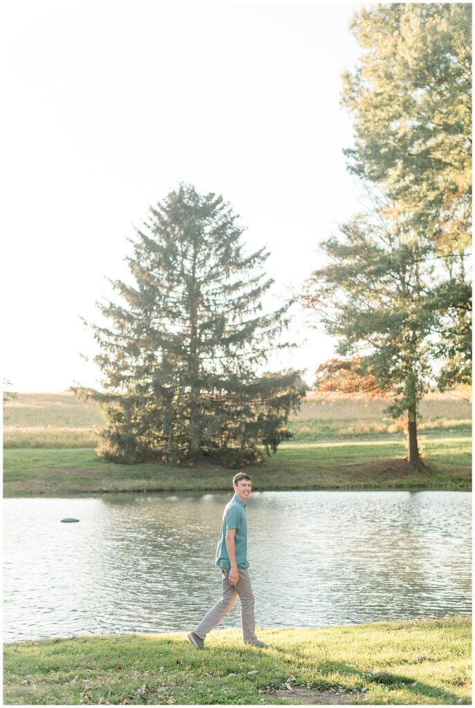 senior guy in teal polo shirt and gray pants walking by pond for this Family Farm Senior Guy Session in Lancaster County