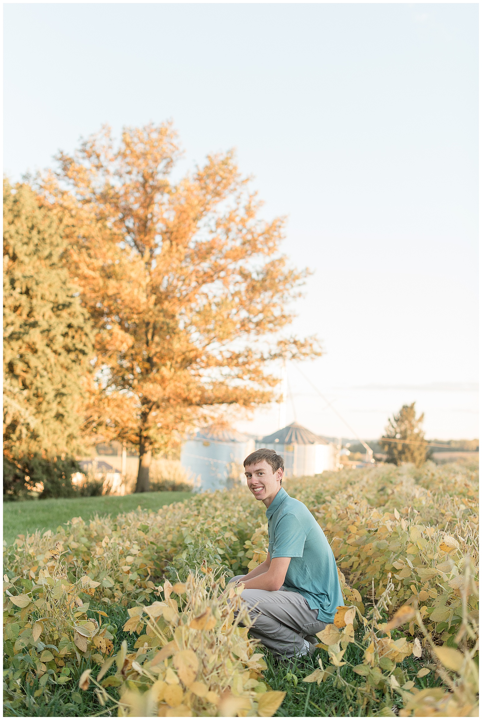 senior guy crouched down in farm field with golden yellow tree in background for this Family Farm Senior Guy Session in Lancaster County