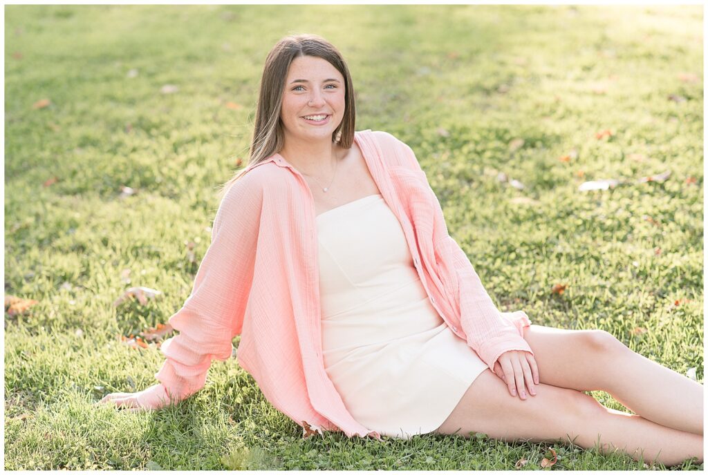 senior girl sitting on grass at sunset in white dress with light pink cardigan for this Longs Park Fall Senior Session