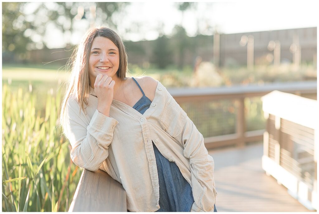 senior girl wearing jean dress and oversized shirt leaning against fence for this Longs Park Fall Senior Session
