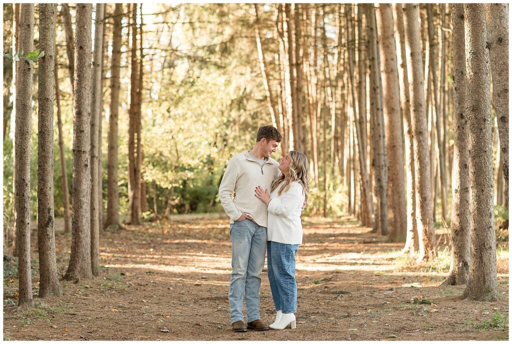 engaged couple in light white shirts and blue jeans standing together by evergreen trees for this Overlook Park Fall Engagement Session